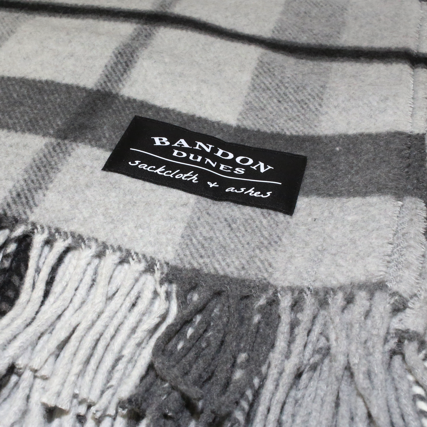 Blanket with gray, charcoal and black plaid design shows fringe and black Bandon Dunes patch.